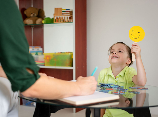 A little girl shows that she is happy while holding a smiley sign to a woman indoors